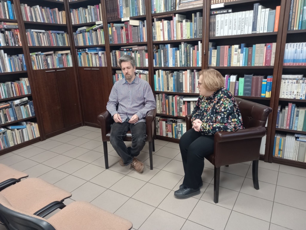 Autor Marcin Mortka rozmawia z Małgorztą Halber, siedzą na fotelach na tle regałów bibliotecznych