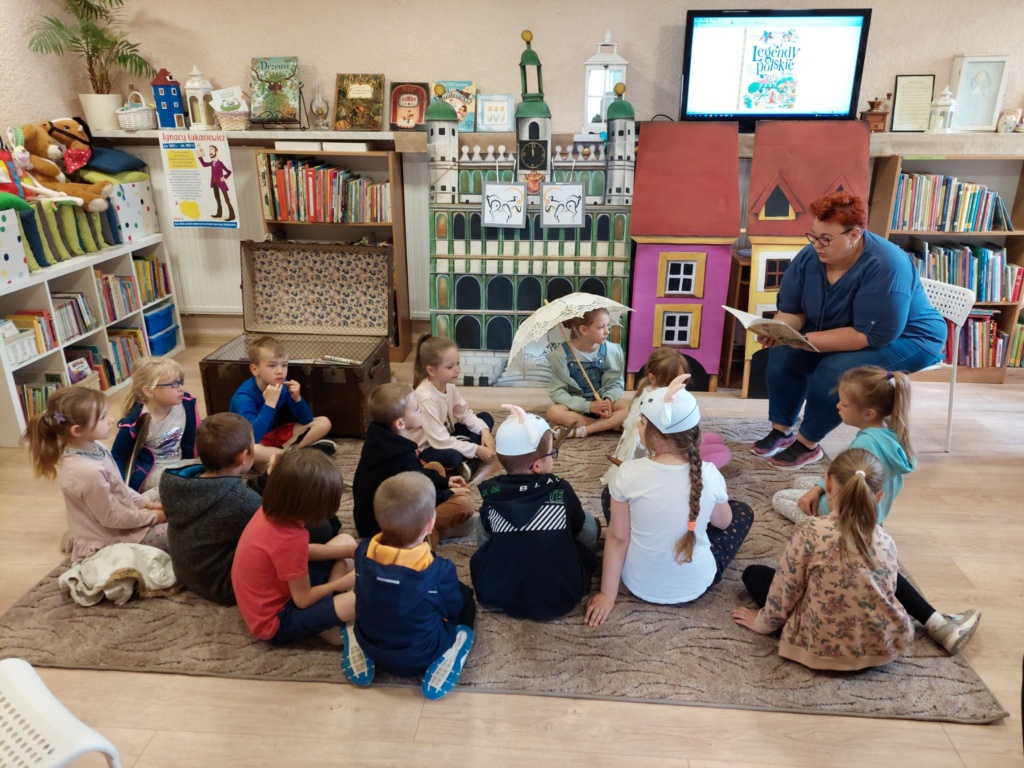 Przedszkolaki siedzą na dywanie i słuchają pani czytającej im ksiązkę