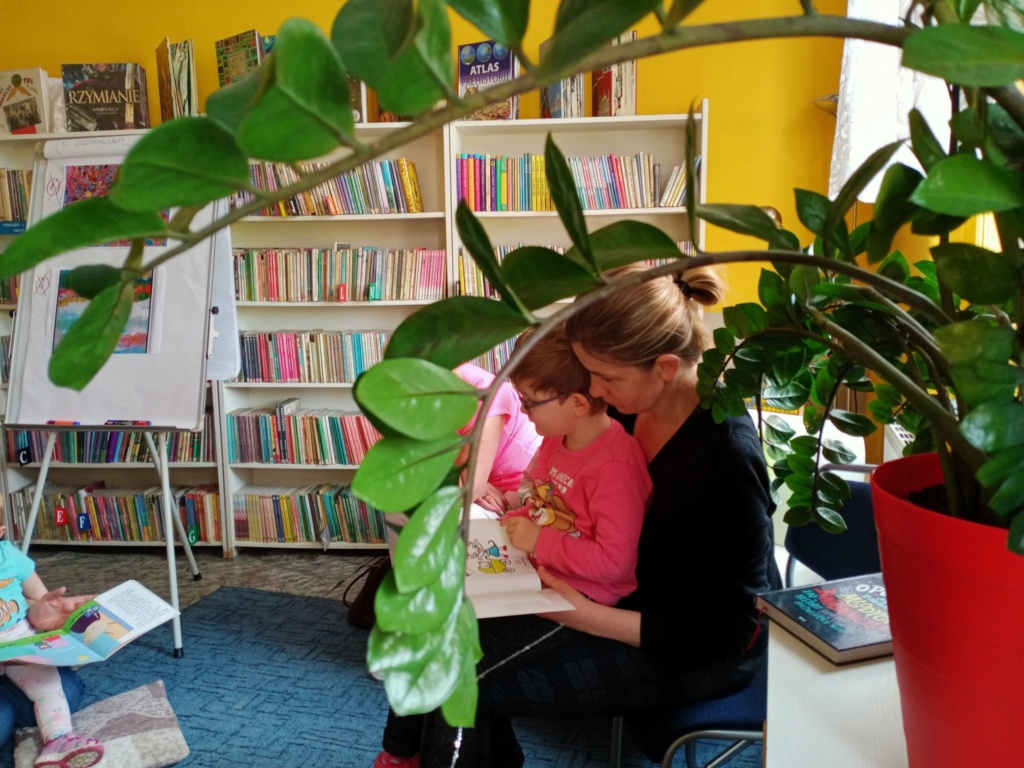 w tle na zdjęciu mama czytająca małemu dziecku książkę w bibliotece, od prawej na czerwonym kole napis głosne czytanie dla najmłodszych